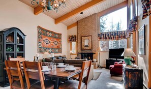 Un salon chaleureux avec une cheminée, un coin repas avec des meubles en bois et une grande fenêtre agrémentée de rideaux.