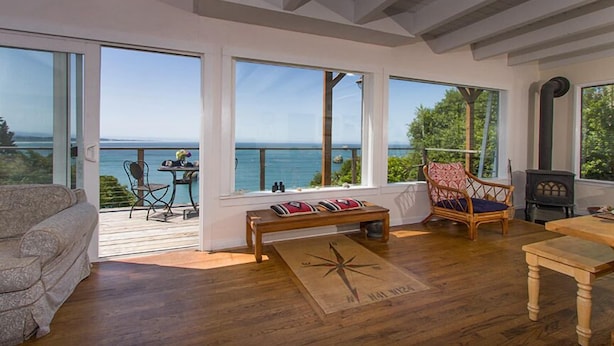 Une pièce spacieuse avec de grandes fenêtres donnant sur la mer, une terrasse en bois et un coin salon confortable avec un banc et des chaises.