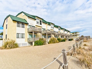 A row of beachfront bungalows with balconies, a sandy walkway, and a wooden fence.