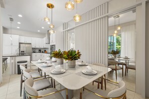 Eine moderne Küche mit weißen Schränken, eine Essecke mit einem weißen Tisch, gedeckt für vier Personen, und Pendelleuchten.