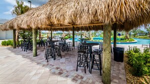 Une terrasse extérieure avec des tables et des chaises surmontées de toits de chaume, une piscine et de la verdure.