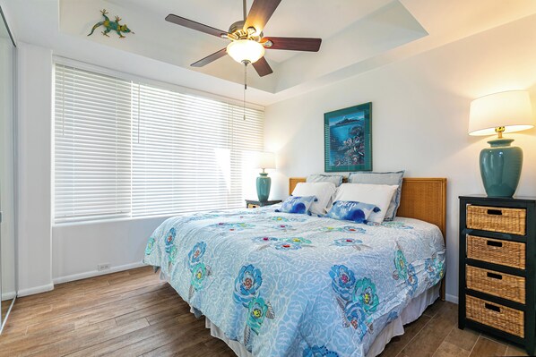A bedroom with a bed, a ceiling fan, a window with blinds, a wall-mounted painting, and a nightstand with a lamp.