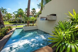 A swimming pool with a waterfall feature, surrounded by palm trees and a well-maintained garden.