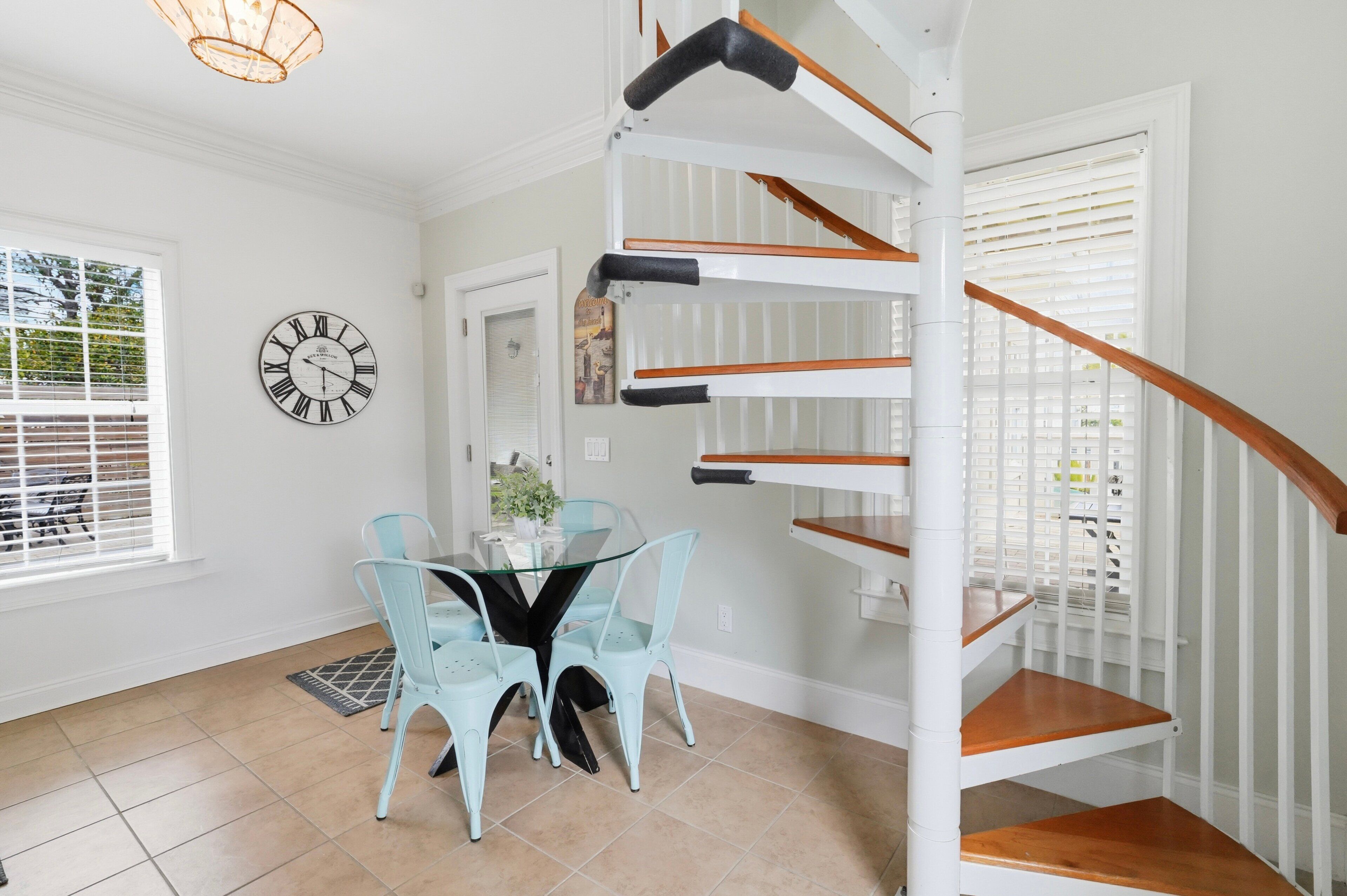 A dining area with a glass table and light blue chairs, a white spiral staircase, and a wall clock.