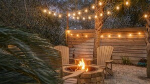 A cozy outdoor seating area with a stone fire pit, wooden chairs, and string lights.