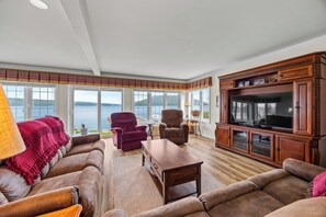 A spacious living room with a large television, comfortable seating, and a view of the water.