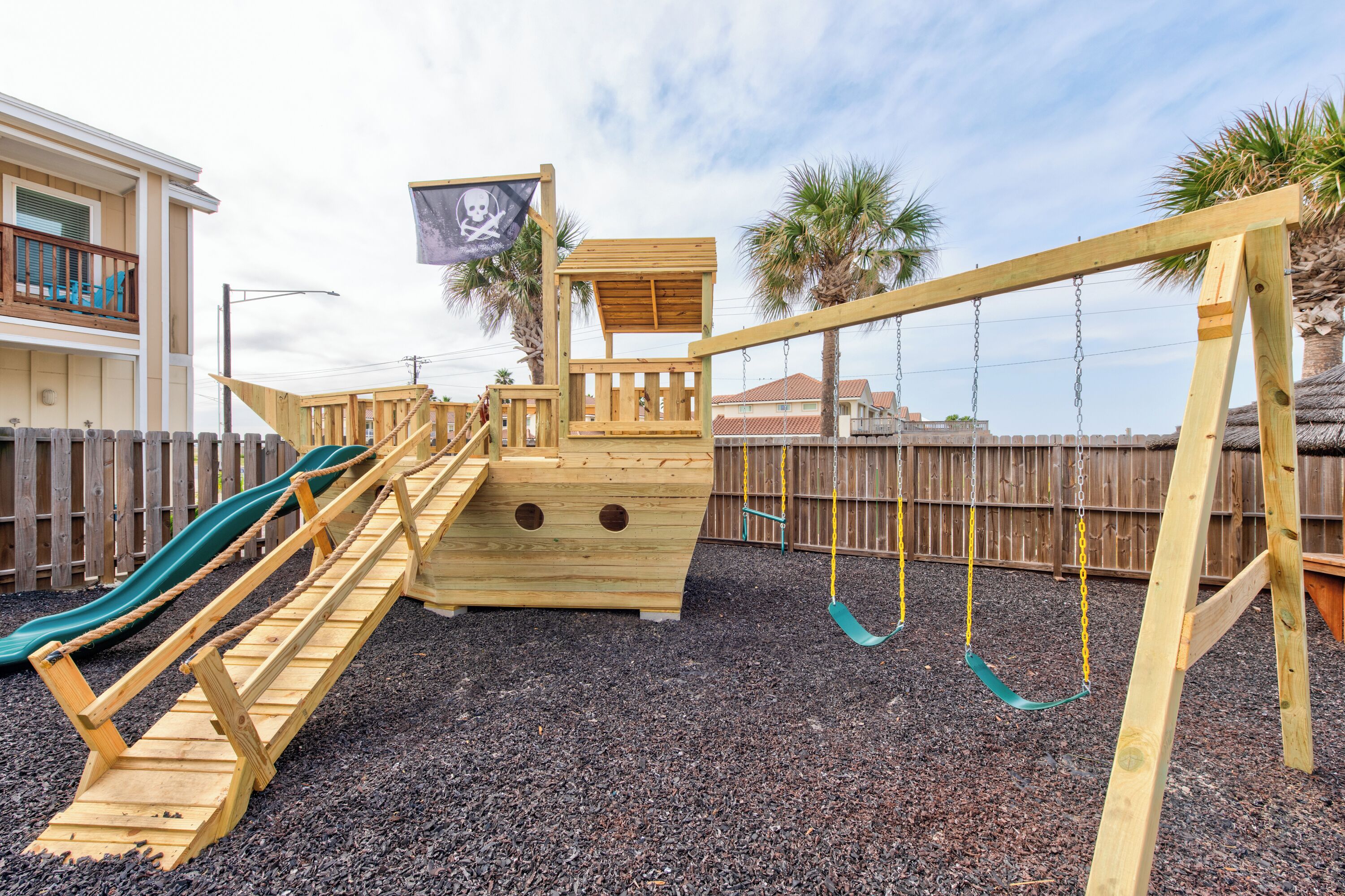 A wooden play structure with a slide, a small pirate-themed playhouse, and a swing set.