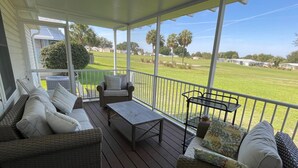 Un porche cubierto con mobiliario de mimbre, una mesa de centro y vistas a un jardín verde y casas a lo lejos.