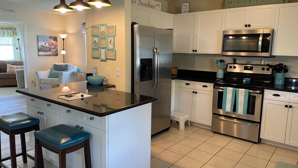 Una cocina moderna con armarios blancos, un refrigerador de acero inoxidable, un horno doble y un microondas. Hay dos taburetes de bar y una pequeña isla con encimera.