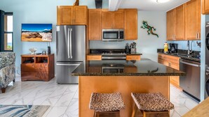 A modern kitchen with wooden cabinets, stainless steel appliances, and a central island with stools. - Maui Vista #1-418 Ocean View/Pickle Ball Courts, Close to the Beach, Wifi (Kihei)