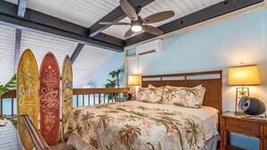 A bedroom with a bed, two wall-mounted fans, a nightstand with a lamp, and three surfboards against the wall. - Maui Vista #1-418 Ocean View/Pickle Ball Courts, Close to the Beach, Wifi (Kihei)