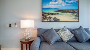 A living room with a grey sofa, a lamp, a small table with a decorative item, and a framed picture of a beach scene on the wall. - Maui Vista #1-418 Ocean View/Pickle Ball Courts, Close to the Beach, Wifi (Kihei)