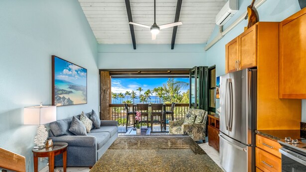 A modern kitchen with a large refrigerator, wooden cabinets, and a dining area with a sofa and table. - Maui Vista #1-418 Ocean View/Pickle Ball Courts, Close to the Beach, Wifi (Kihei)