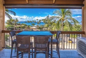 Ein Balkon mit Korbstühlen und einem Tisch mit Blick auf einen tropischen Strand, an dem Palmen stehen und ein klarer, blauer Himmel zu sehen ist.
