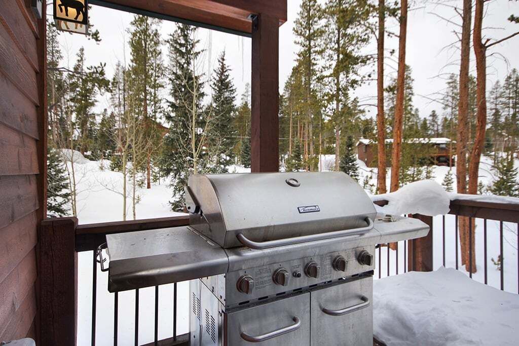 Una parrilla para barbacoa en una terraza exterior con vista a árboles nevados y una cabaña al fondo.