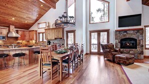 A spacious kitchen with wooden cabinets, a large island with stools, a stone fireplace, and a dining area with chairs.