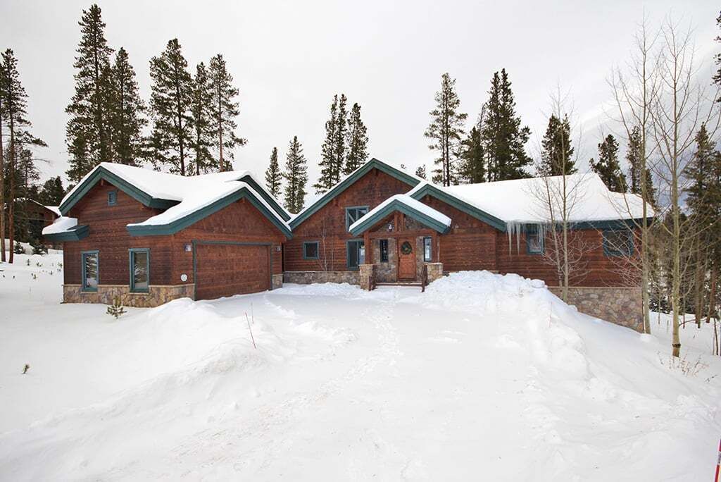 A log cabin with a snow-covered roof and a stone foundation, surrounded by snow and tall pine trees.