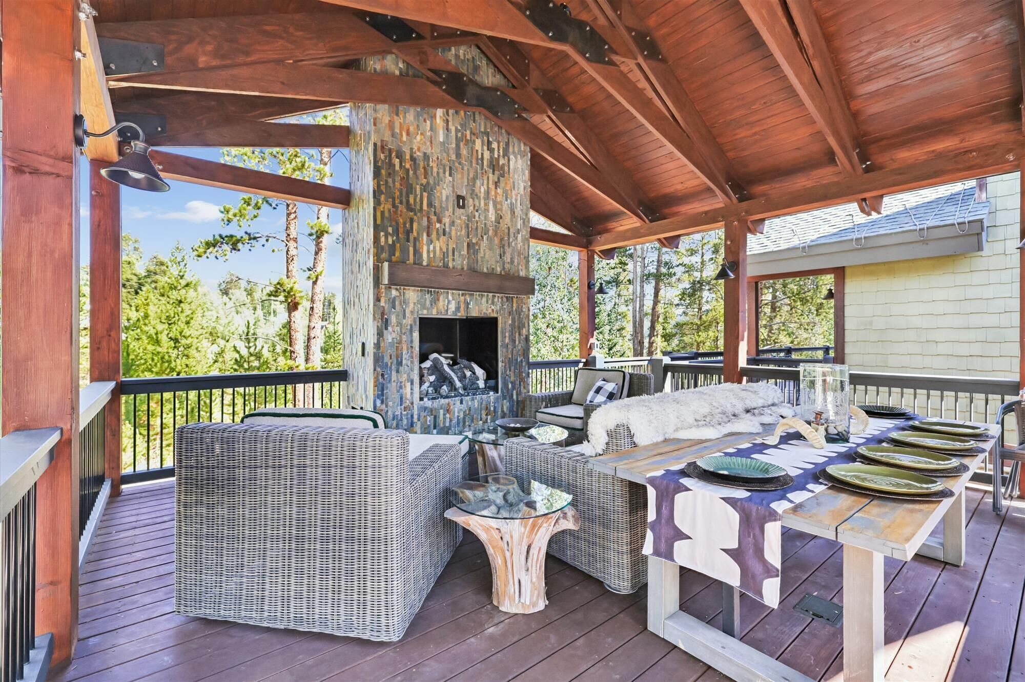 A covered outdoor patio with wicker furniture, a stone fireplace, and a dining table set for a meal.