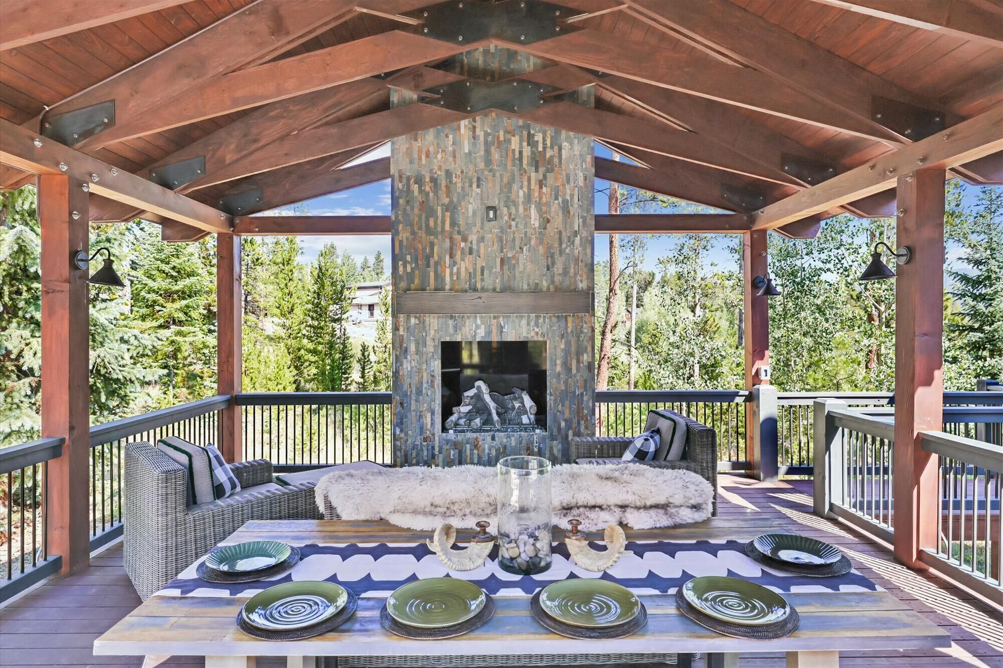 A covered outdoor seating area with a stone fireplace, wicker furniture, and a view of trees.