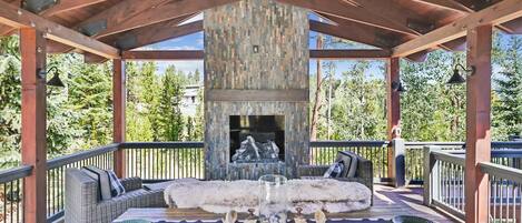 A covered outdoor seating area with a stone fireplace, wicker furniture, and a view of trees.