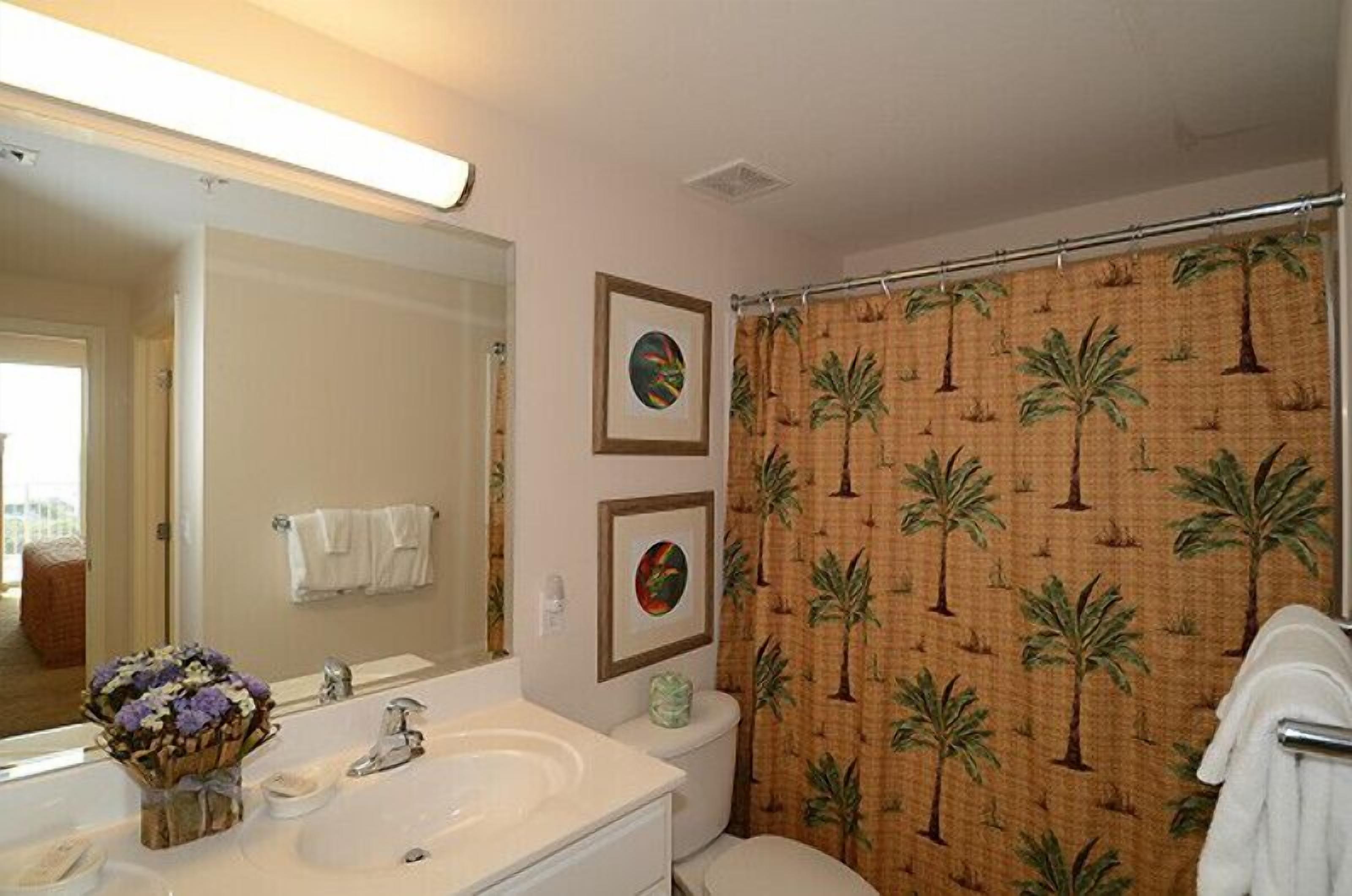 Een badkamer met een witte wastafel, een toilet en een douchegordijn met palmboommotieven. Twee ingelijste foto's hangen aan de muur en er staat een vaas met bloemen op de wastafel.