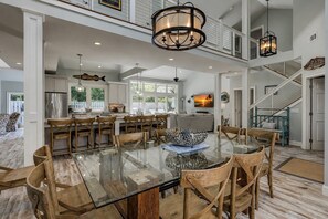 A spacious dining area with a glass-top table surrounded by wooden chairs, a kitchen in the background, and a staircase leading to an upper level.