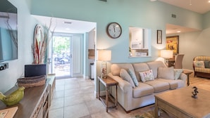A living room with a beige sofa, a coffee table, a TV, and a dining area with a chair and table. - Charming 2BR Beach Villa! GOLF CART, Tram, Beach, Pool, Porch, Near Baytowne. (Destin)