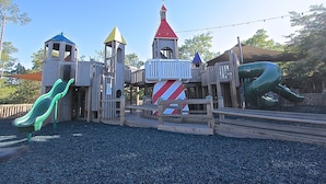 A wooden play structure with slides and climbing areas, surrounded by gravel. - Charming 2BR Beach Villa! GOLF CART, Tram, Beach, Pool, Porch, Near Baytowne. (Destin)