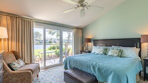 A hotel room with a bed, a chair, a table, and a view of the outdoors. - Charming 2BR Beach Villa! GOLF CART, Tram, Beach, Pool, Porch, Near Baytowne. (Destin)