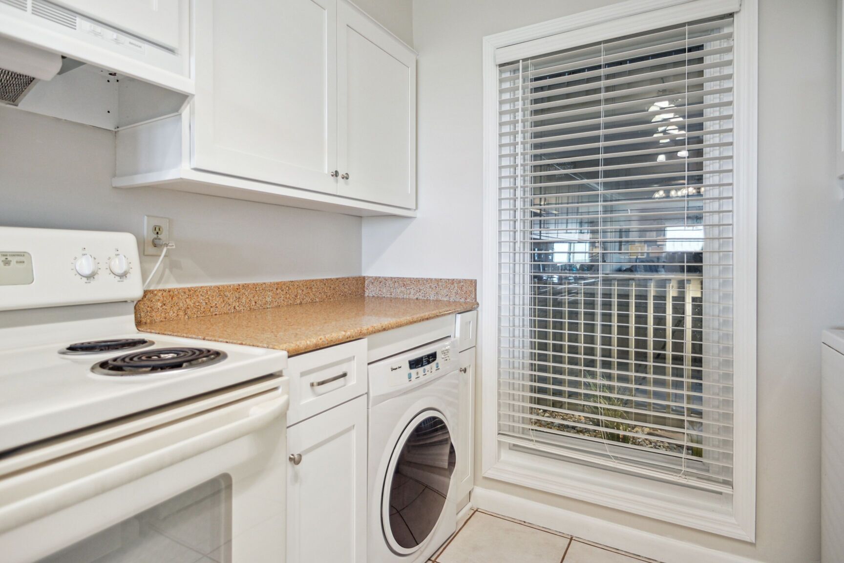 A kitchen with white cabinets, a granite countertop, a stove, a washing machine, and a window with blinds.