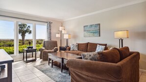 A modern living room with a sofa set, a coffee table, and a side table. There is a large window with a view of greenery and a palm tree. - Shorewood 103 - Oceanfront 1st Floor Condo (Hilton Head Island)