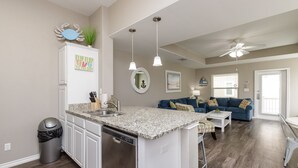 A modern kitchen with a central island, a dining area with a white table and blue sofa, and a living room with a blue sofa and a ceiling fan. - Pet-Friendly Beach Retreat | Heated Pool & Walk to Beach (Corpus Christi)