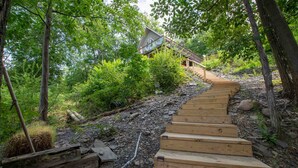 A wooden staircase leading up to a house surrounded by lush greenery. - All Decked Out: Beautiful Chalet-style Seneca Lake Home! (Burdett)