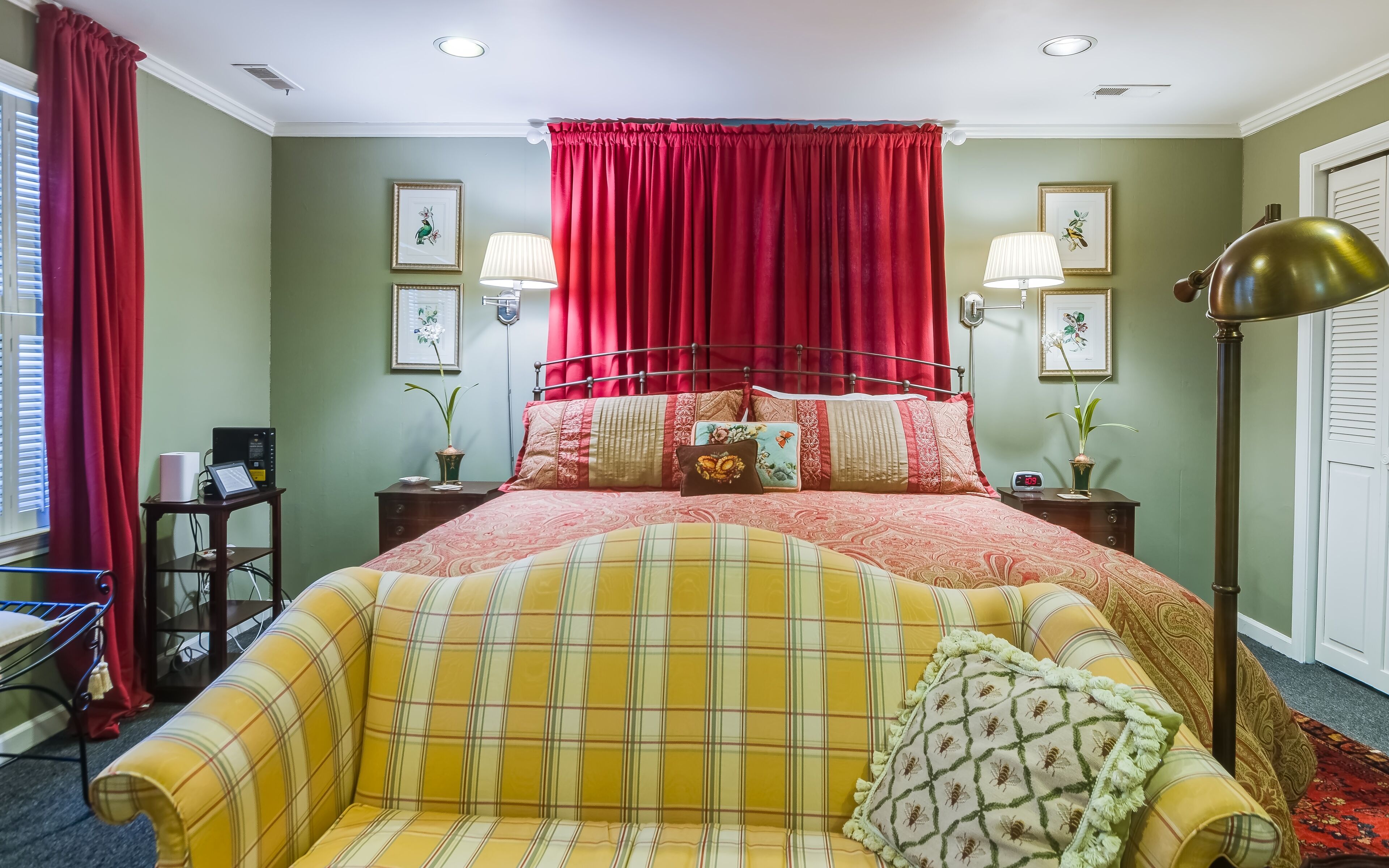 Een slaapkamer met een bed, een nachtkastje met een lamp, een gordijn in rood en een raam met zonwering.