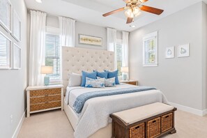 Un dormitorio con cabecera capitonada, cama con ropa de cama azul y blanca, mesita de noche con lámpara y ventilador de techo.