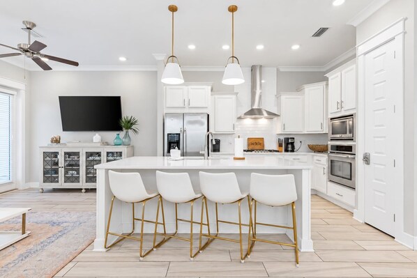 Una cocina moderna con armarios blancos, una isla central con taburetes y un ventilador de techo, iluminación empotrada y un televisor de pantalla plana.