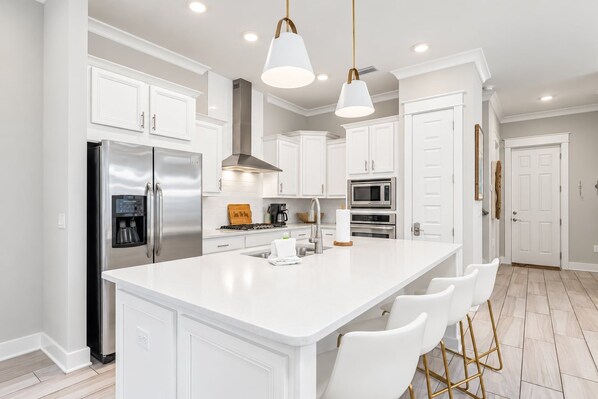 Cocina moderna con armarios blancos, electrodomésticos de acero inoxidable, una isla central con taburetes y luminarias colgantes.