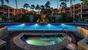 A swimming pool with a hot tub, surrounded by palm trees and lounge chairs, with a hotel building in the background.