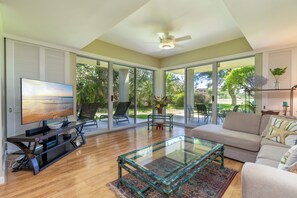 A spacious living room with a television, a glass coffee table, a sofa, and a ceiling fan.