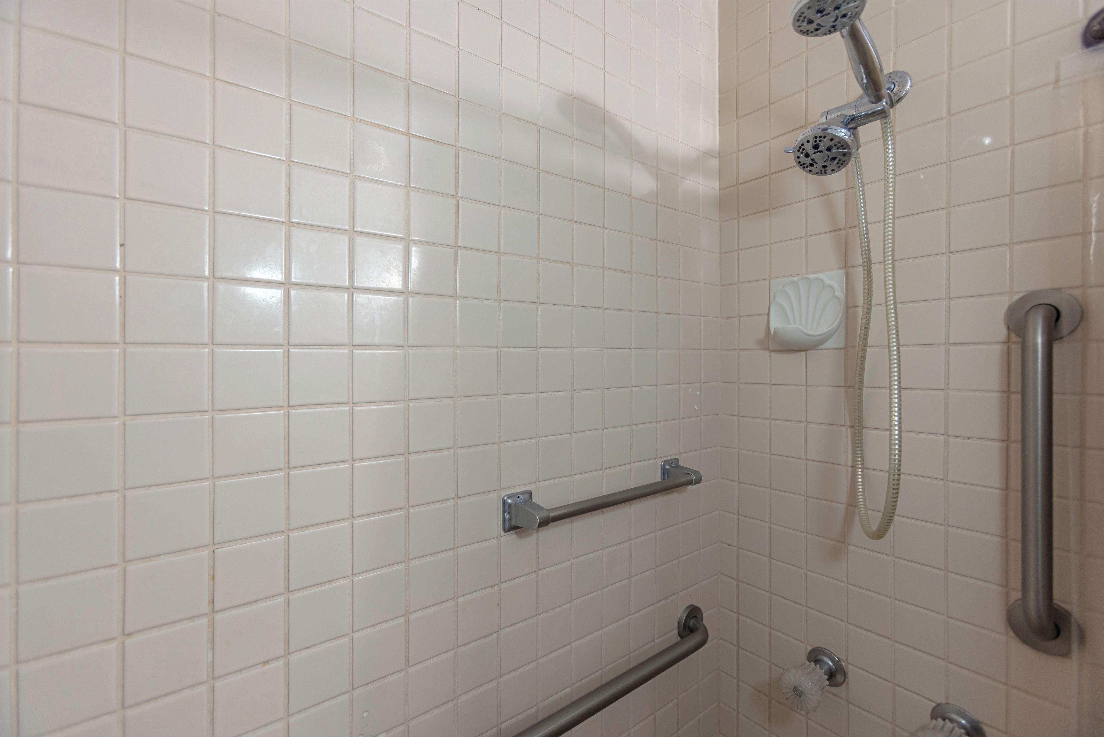 Une salle de bain avec des carreaux blancs, une douchette à main, un flexible de douche, une barre d’appui et un porte-savon.