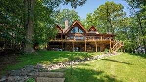 A large, two-story log cabin with a deck and stone foundation, surrounded by lush greenery.