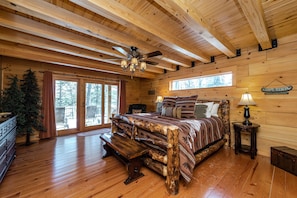 A log cabin bedroom with a wooden bed, a ceiling fan, and a large window.
