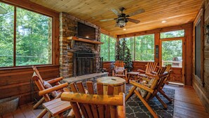 A log cabin interior with a stone fireplace, wooden furniture, and large windows offering a view of the forest.