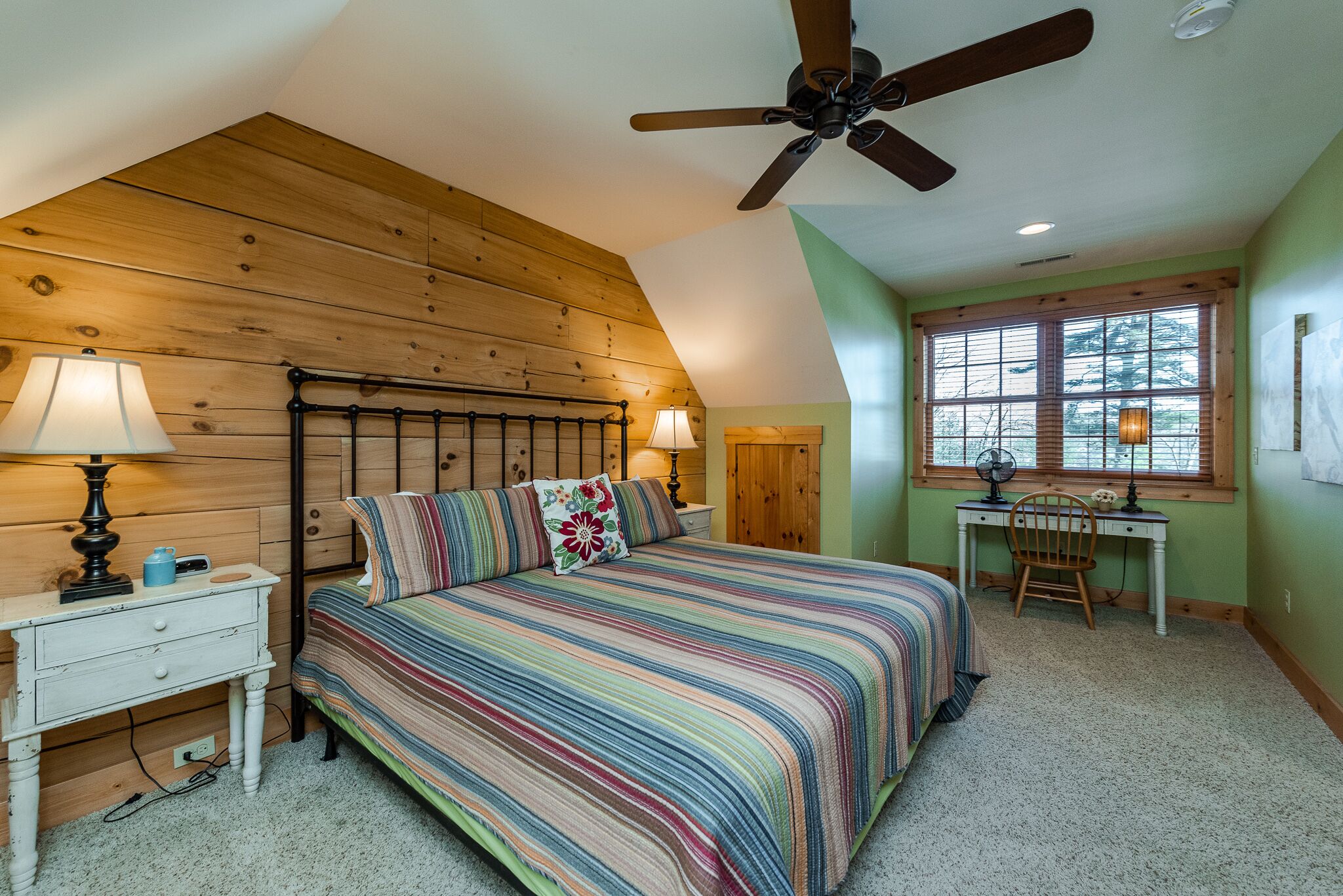 Un dormitorio con techo de madera, una cama con colcha a rayas, dos lámparas de noche, un escritorio con silla y una ventana con vistas a árboles.