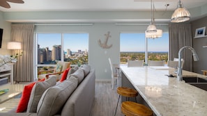 Enjoy a panoramic city view from this spacious living area with a sofa, chairs, and kitchen with a large window.