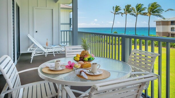 A balcony with white chairs and a table set for breakfast, overlooking a clear blue ocean and palm trees. - Poipu Sands 424 Exquisite Oceanview  By Suite Paradise (Koloa)