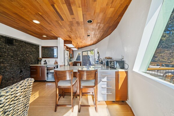 A modern kitchen with a stone wall, wooden ceiling, and large windows. - Dramatic Geodesic Dome with amazing views, AC, spa and walk to town cold tub (Idyllwild)
