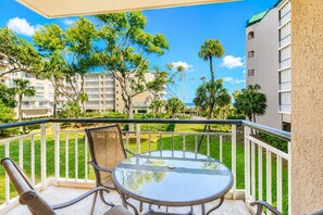 A balcony with a glass table and chairs, overlooking a well-maintained lawn and buildings.
