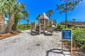 A playground with a slide and stairs, surrounded by sand and greenery, with a 'Welcome' sign.