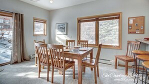A dining area with a wooden table and chairs, large windows with blinds, a framed picture on the wall, and a bulletin board with notices. - Affordable Ski In/Out Mountain Oasis: Recent Updates, Easy Walk to Main St (Breckenridge)